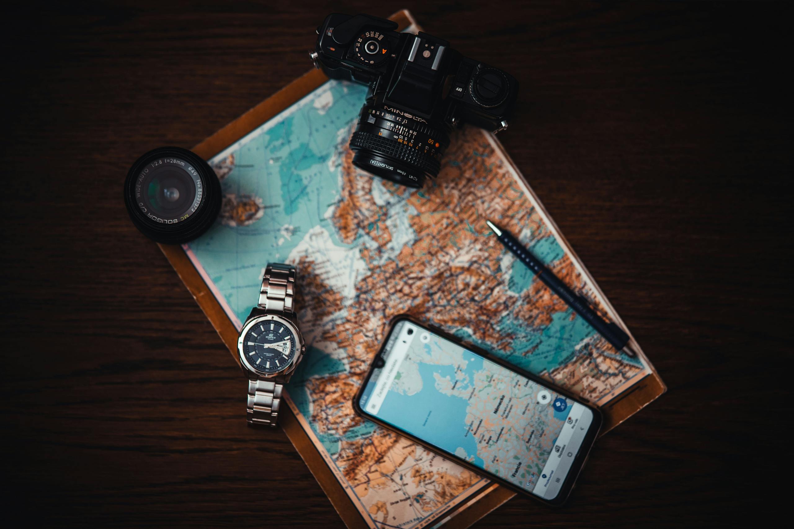 Top view of travel essentials including a map, camera, phone, watch, and lens on a wooden surface.